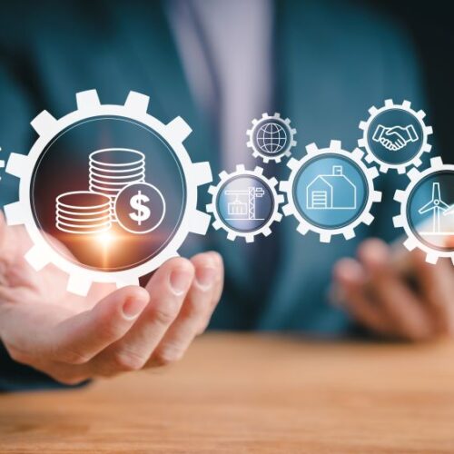 Hand holding a gear with money coins, surrounded by icons for industry, construction, energy, and global collaboration
