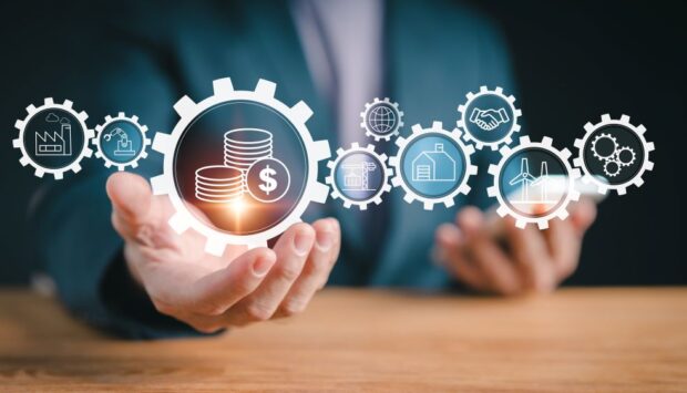 Hand holding a gear with money coins, surrounded by icons for industry, construction, energy, and global collaboration